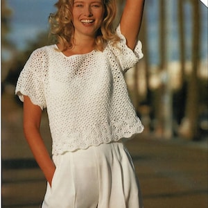 Op de afbeelding: Een wit gehaakt topje met korte mouwen en een gegolfde zoom. Het topje is gemaakt met een delicaat open steekpatroon.