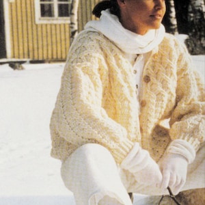 Puede incluir: Un cárdigan blanco de punto de cable con una bufanda blanca y pantalones blancos. La persona está sentada en un entorno exterior nevado.