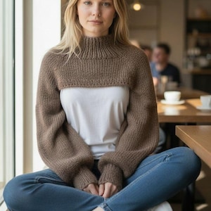 May include: A brown, high-neck knitted shrug with long sleeves. The cropped sweater is worn over a white t-shirt and paired with blue jeans and white sneakers. The model is sitting in a cafe setting.