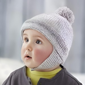 May include: A baby wearing a grey knitted hat with ear flaps and a pom-pom on top. The hat has a white and grey striped pattern. The baby is wearing a grey shirt with yellow underneath.