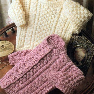 Könnte beinhalten: Zwei gestrickte Baby-Pullover, einer weiß und einer rosa, mit einem Zopfmuster. Der rosa Pullover hat Knöpfe vorne.