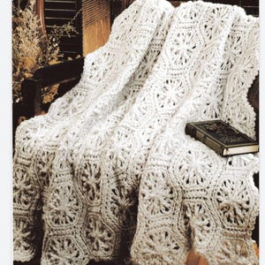 May include: A cream-colored, crocheted blanket with a floral pattern draped over a wooden chair. The blanket has a textured, open weave design. A small, dark book rests on the blanket.