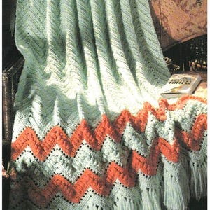 May include: A crocheted throw blanket with a green and orange chevron pattern. The blanket has a fringe border and is draped over a chair.