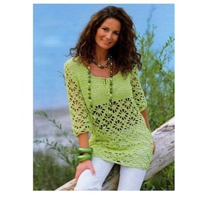 May include: A light green crochet tunic with three-quarter sleeves and a floral pattern. The tunic is paired with white pants and green bracelets. A beaded necklace complements the outfit. The background features a blue sky and green foliage.