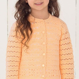 Peut inclure: Un cardigan pêche en crochet avec un col rond et une fermeture boutonnée. Les manches et le corps présentent un motif ajouré. Le cardigan est porté sur une robe blanche. L'image montre une jeune fille aux longs cheveux bruns ondulés.