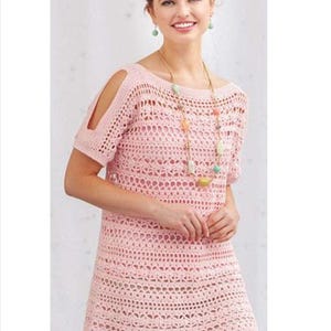 Op de afbeelding: Een lichtroze gehaakte tuniek met korte mouwen en schouderuitsparingen. De tuniek heeft een open, kantachtig ontwerp en een ronde halslijn. Een lange kralenketting met kleurrijke kralen is ook zichtbaar.