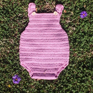 以下が含まれることがあります： ピンクのクロシェで編まれたベビーロンパース。ストラップにボタン留めがあります。ロンパースは緑の芝生の上に置かれています。