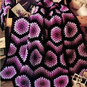 Puede incluir: Una manta de ganchillo hecha a mano con un patrón floral repetido en tonos morado, rosa y blanco sobre un fondo negro. La manta está sobre una superficie de madera, con fotografías antiguas y otros objetos visibles.