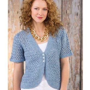 May include: A light blue, short-sleeved crochet cardigan with a V-neck and three white buttons. The cardigan is worn over a white top and paired with a gold chain necklace. The background is a wooden wall.