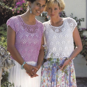 May include: Two women wearing crocheted tops with a lacy pattern. The woman on the left is wearing a light purple top and the woman on the right is wearing a white top. Both women are wearing skirts.