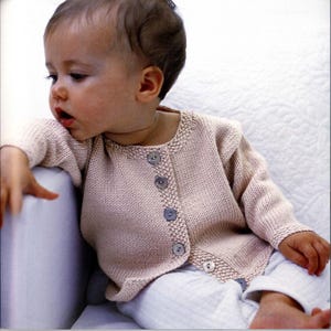 May include: A baby wearing a light pink knitted cardigan with button closures. The cardigan has a simple design and is made with a soft, textured yarn.