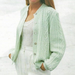 May include: A woman wearing a light green cable knit cardigan with white buttons over a white lace tank top and white pants.