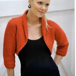 Puede incluir: Un bolero de crochet rojo con cuello de chal. El bolero se lleva sobre un top negro de tirantes.