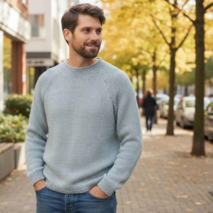 May include: A pale blue knitted jumper with a crew neck and long sleeves. The jumper is made of a soft, textured material. The person is wearing blue jeans and standing on a pavement with trees in the background.