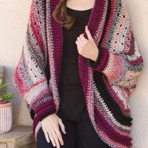 May include: A crocheted cardigan with a wide, open collar and long sleeves. The cardigan is made with a multi-coloured yarn in shades of pink, black, and grey.