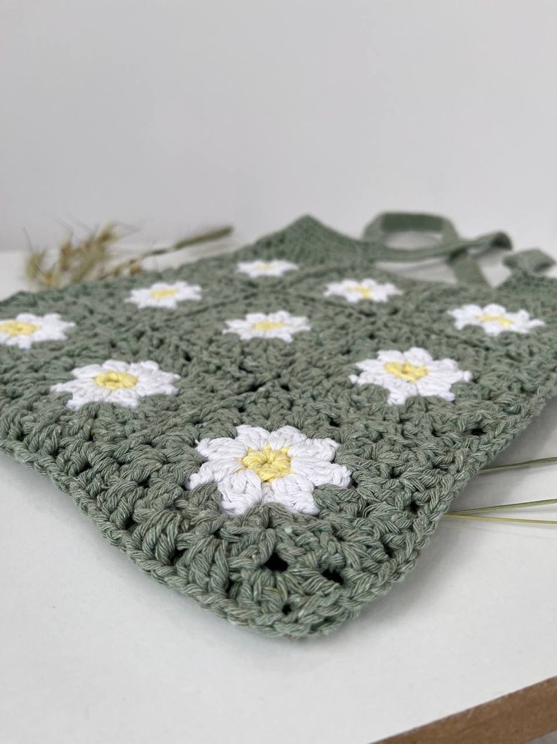 Peut inclure: Un sac en crochet vert avec des fleurs de marguerite blanches et jaunes.