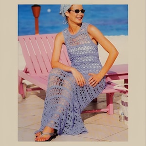 Puede incluir: Una mujer lleva un vestido largo de crochet de color lila claro con un sombrero de crochet blanco y gafas de sol. Está sentada en una silla de madera rosa en una playa.