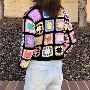 May include: A black crochet cardigan with a colorful patchwork design. The cardigan is made up of squares in various shades of pink, blue, yellow, green, and brown. The cardigan is cropped and has a relaxed fit.