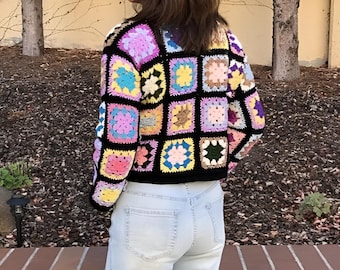 Könnte beinhalten: Eine schwarze Häkeljacke mit einem bunten Patchwork-Muster. Die Jacke besteht aus Quadraten in verschiedenen Farbtönen von Rosa, Blau, Gelb, Grün und Braun. Die Jacke ist kurz geschnitten und hat eine lockere Passform.