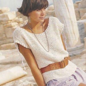 Peut inclure: Un haut en crochet blanc avec un col rond et des manches courtes. Le haut est doté d'une ceinture en cuir marron avec une boucle argentée. Le mannequin porte le haut avec un collier en chaîne argentée et des boucles d'oreilles.