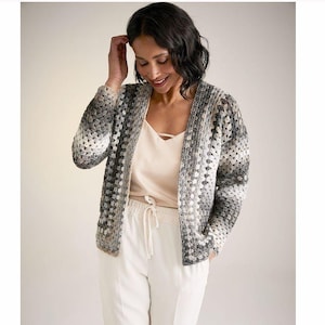 May include: A grey, white and black crocheted cardigan with a long sleeve and open front. The cardigan is worn over a white vest top and white trousers.