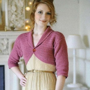 May include: A pink crocheted bolero with a button fastening. The bolero has a collar and short sleeves. The model is wearing the bolero over a cream-coloured dress.