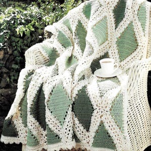 May include: A crocheted throw blanket with a diamond pattern in shades of green and white. The blanket is draped over a white wicker chair with a cup of tea on a saucer.