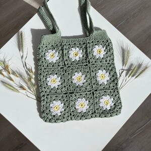 Peut inclure: Un sac cabas en crochet vert avec des fleurs de marguerite blanches. Le sac a deux anses et est fait d'un fil doux et textur&eacute;.