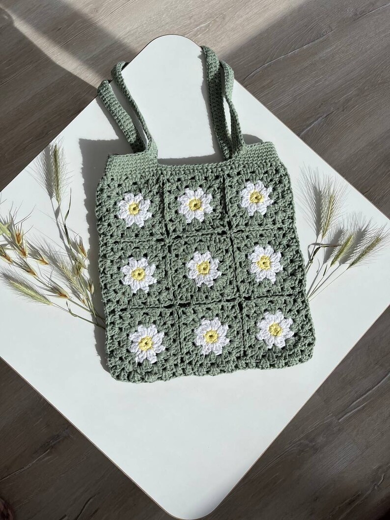 Peut inclure: Un sac cabas en crochet vert avec des fleurs de marguerite blanches. Le sac a deux anses et est fait d'un fil doux et textur&eacute;.