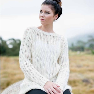 Könnte beinhalten: Cremefarbener Strickpullover mit offenem Strickmuster, langen Ärmeln und einem U-Boot-Ausschnitt. Der Pullover hat ein strukturiertes Muster. Die Person trägt schwarze Leggings. Der Hintergrund zeigt ein Feld und einen bewölkten Himmel.