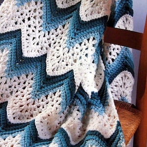 Op de afbeelding: Een gehaakt deken met een chevron-patroon in tinten blauw en wit. Het deken is over een houten stoel gelegd.