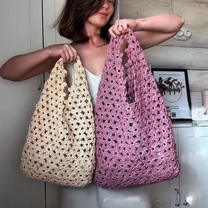 Op de afbeelding: Twee gehaakte draagtassen, één crème en één roze, worden getoond. De tassen zijn gemaakt van een bloemenpatroon. De tassen hebben een grote opening en een handvat. De achtergrond is een lichtgekleurde muur.