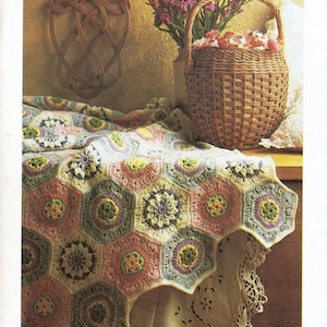 Puede incluir: Una manta de ganchillo con un patrón hexagonal en tonos de rosa, verde, amarillo y blanco. La manta está drapeada sobre una mesa con un mantel de encaje y una cesta de mimbre llena de flores.