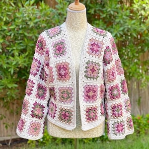 May include: A white crocheted cardigan with a pink and green granny square pattern. The cardigan has a V-neck and long sleeves.
