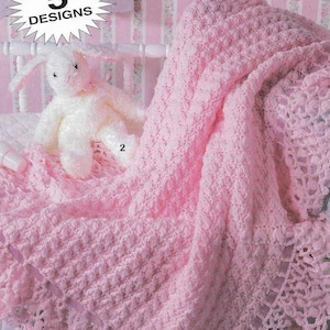 May include: A pink crocheted baby blanket with a lacy border. The blanket is draped over a white crib with a white stuffed bunny sitting on it. The blanket has a textured pattern and is made with a soft yarn. The image includes the text "5 DESIGNS".