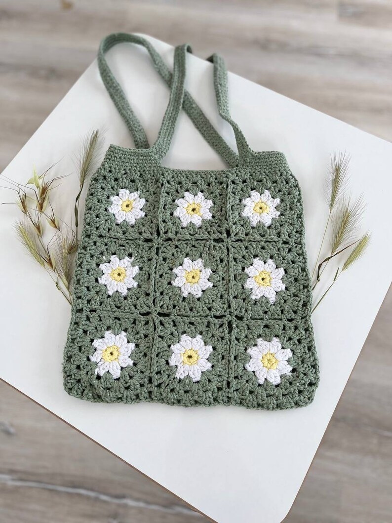 Peut inclure: Un sac cabas en crochet vert avec des fleurs de marguerite blanches et jaunes. Le sac a deux longues anses.