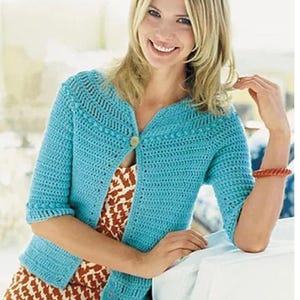 May include: A turquoise crochet cardigan with a button fastening. The cardigan features short sleeves and a decorative collar. The woman is wearing a patterned dress and a red bracelet.