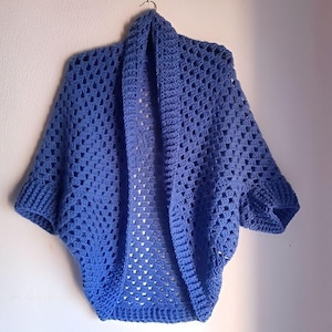 May include: A light blue crocheted shrug with short sleeves. The shrug is hanging on a hanger against a white wall.