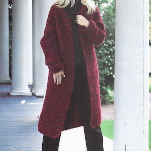 May include: A long, burgundy knitted coat with a textured finish. The coat is open at the front and reaches the ankles. The person is wearing black trousers and shoes. The background features white columns and a blue floor.