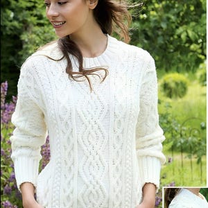 Puede incluir: Un suéter blanco de punto de cable con cuello redondo y mangas largas. El suéter tiene un patrón de punto de cable detallado.