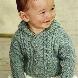 May include: A baby boy wearing a light green knitted sweater with a cable knit pattern and a hood. The sweater has a relaxed fit and is perfect for a casual look.