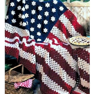May include: A crocheted blanket in the colors of the American flag. The blanket is folded over a wooden bench and has a red, white, and blue color scheme.