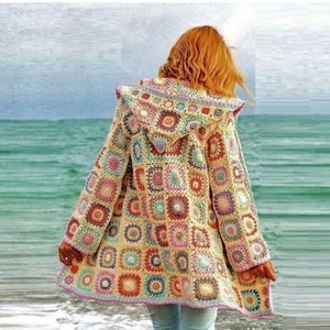 May include: A long, crocheted cardigan with a hood. The cardigan is made of colourful squares and has a light beige background. The cardigan is being worn by a person standing on a beach.