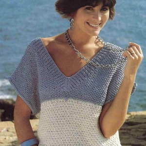 May include: A woman wearing a blue and white knitted top with a v-neck and short sleeves. She is wearing a silver chain necklace and a blue bracelet.