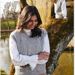 Puede incluir: Un chaleco de punto gris con cable y cuello en V. El chaleco se lleva sobre una blusa blanca y pantalones marrones.