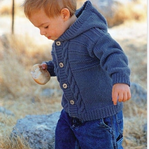 May include: A young child wearing a blue knitted hooded cardigan with a cable knit pattern and button closure. The cardigan has a hood and is worn over a pair of blue jeans.