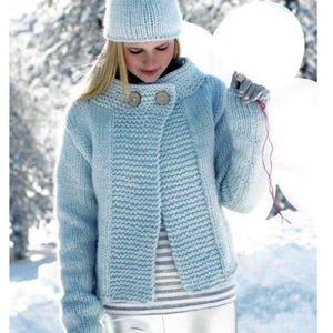 May include: A light blue knitted cardigan with a button closure and a matching light blue knitted hat. The cardigan has a slightly oversized fit and a relaxed silhouette. The model is wearing the cardigan with silver pants and a striped shirt.