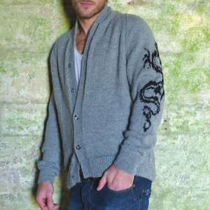 Könnte beinhalten: Grauer Cardigan mit einem schwarzen Drachendesign auf dem linken Ärmel. Der Cardigan hat einen Schalkragen und einen Knopfverschluss. Der Text "Dragon Cardigan" befindet sich am unteren Bildrand. Die Person trägt eine blaue Jeans.
