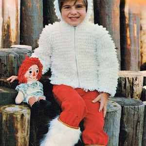 May include: A child wearing a white, textured, hooded jacket with a zipper, paired with red pants and white furry boots. The child is holding a Raggedy Ann doll. The background features wooden posts.