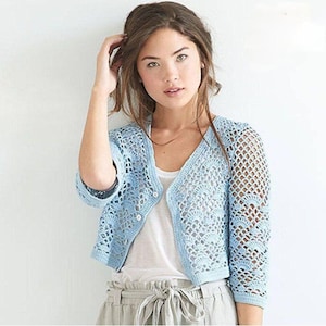 Puede incluir: Un cárdigan de crochet azul claro con cierre de botón y un delicado patrón de calado. El cárdigan es corto y tiene mangas tres cuartos.